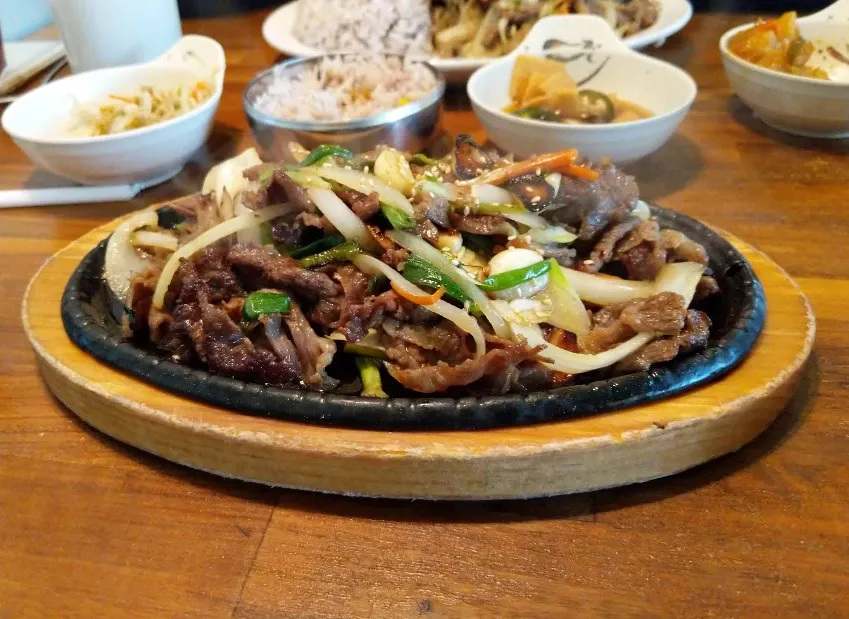 Placeholder photo: sizzling sliced beef with onions and scallions on a hot plate, with side dishes behind