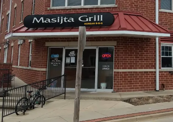 Placeholder photo: the Masijta Grill storefront on a brick building with an open sign, shown blurred